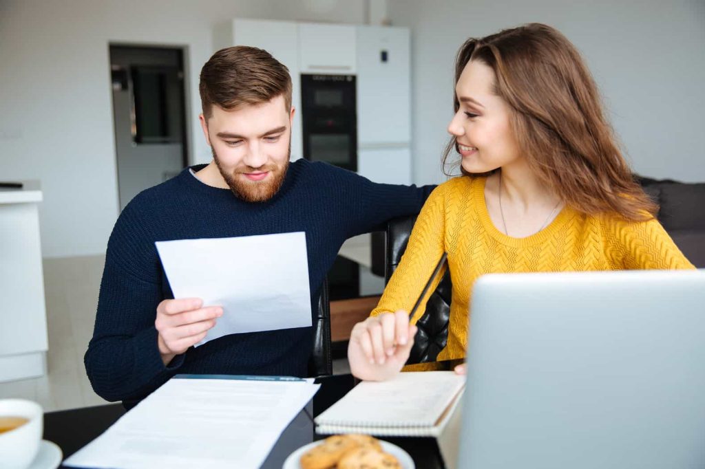 A man and a woman are sitting at a table. The man, holding a piece of paper, and the woman, smiling and holding a notebook, are engaging in a conversation. On the table are a laptop, some documents, a white cup, and a plate of cookies.