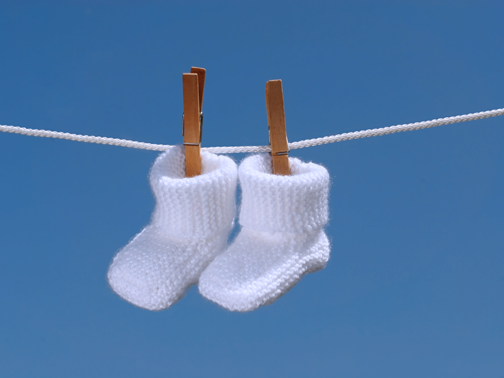 Two white baby booties are clipped to a clothesline with wooden clothespins against a bright blue sky. The knitted booties appear soft and cozy, suggesting a serene and gentle atmosphere.