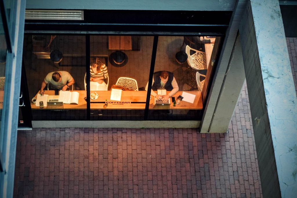 Overhead view through a large window of three people sitting at a counter inside a coffee shop, each working on their laptops or reading books. The coffee shop is lit warmly, contrasting with the outdoor brick path seen through the window.
