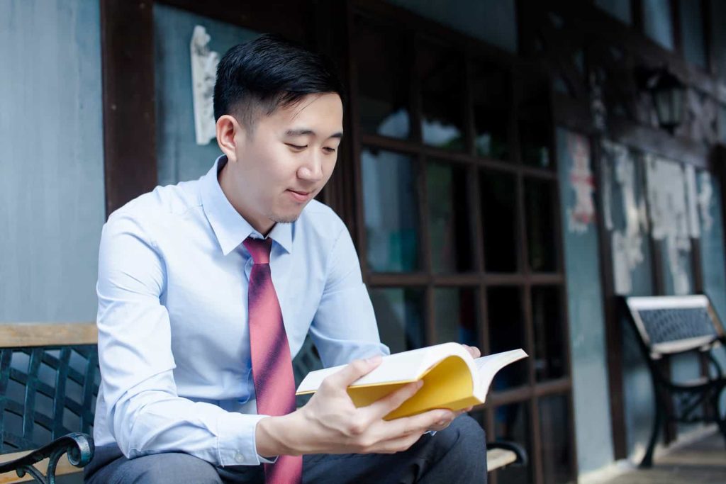 A young man in a light blue shirt and red tie sits on a bench, holding an open book and reading. He appears focused and is outdoors, with a building facade in the background featuring large windows and wooden elements.