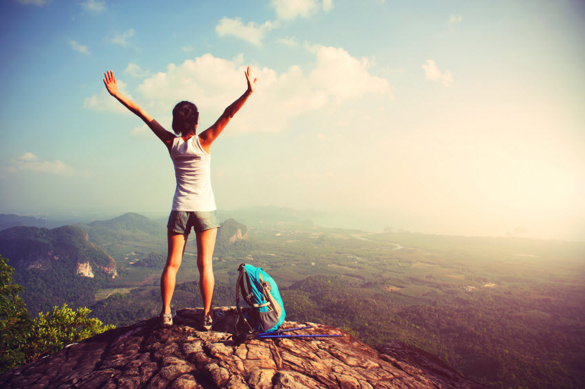 A person with a backpack stands on a rocky cliff with arms raised, overlooking a vast, scenic landscape of mountains, valleys, and rivers under a bright, cloudy sky. The scene captures a sense of freedom and adventure.