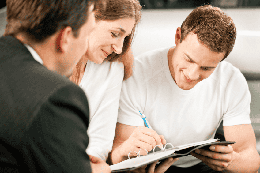 Three people are seated and engaged in a discussion. A man in a white shirt is signing a document on a clipboard, while a woman beside him and another man in a suit attentively watch. They appear to be in a professional setting, possibly finalizing a deal.