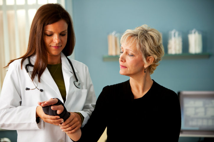 A female doctor in a white coat examines a wrist brace on an older female patient's wrist. The doctor has a stethoscope around her neck, and the patient, dressed in black, looks down at her wrist.