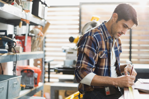 A man in a plaid shirt works at a desk in a workshop filled with tools and equipment. Shelves with various items are visible, and a table saw is in the background. He is focused, marking measurements on a piece of wood.