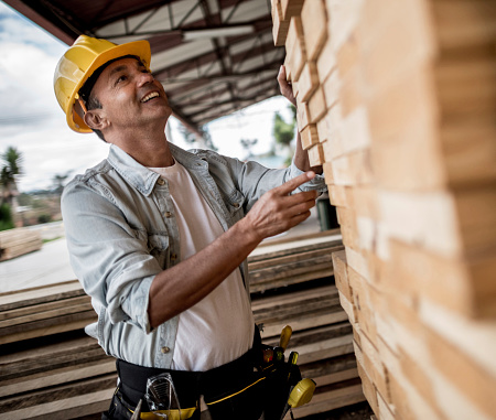 A construction worker wearing a yellow hard hat and a tool belt examines stacked wooden planks at an outdoor lumberyard. He is smiling and looking up at the planks under a partially covered roof.