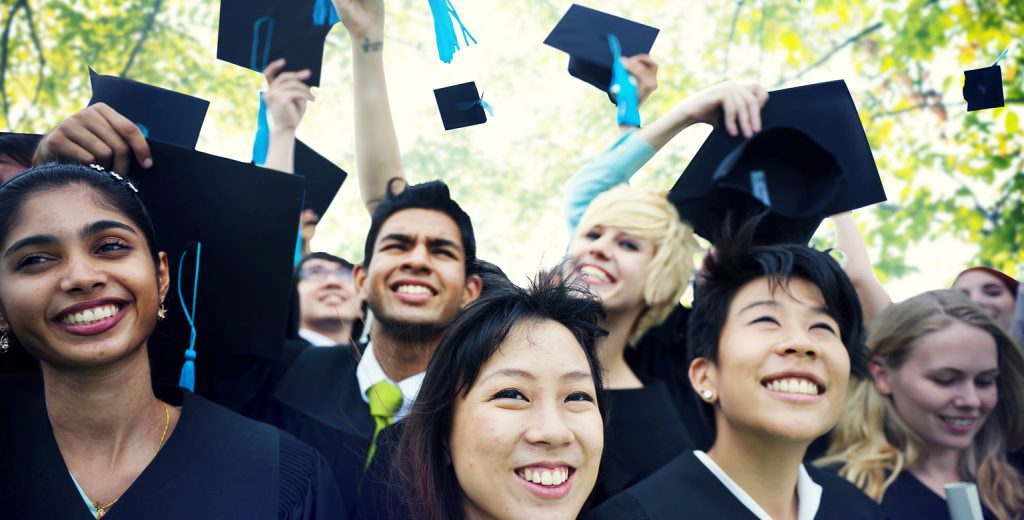 A joyful group of graduates in caps and gowns celebrate by throwing their mortarboards into the air. They are standing outdoors on a sunny day, smiling and looking upwards, with trees and sunlight filtering through the leaves in the background.
