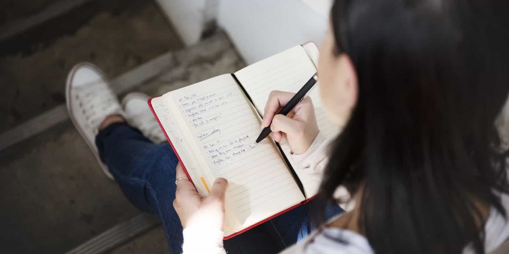 A person with long dark hair, wearing a light-colored sweater and blue jeans, is sitting on steps and writing in an open notebook with lined pages. The person holds a pen in their right hand and appears to be making notes or a journal entry.
