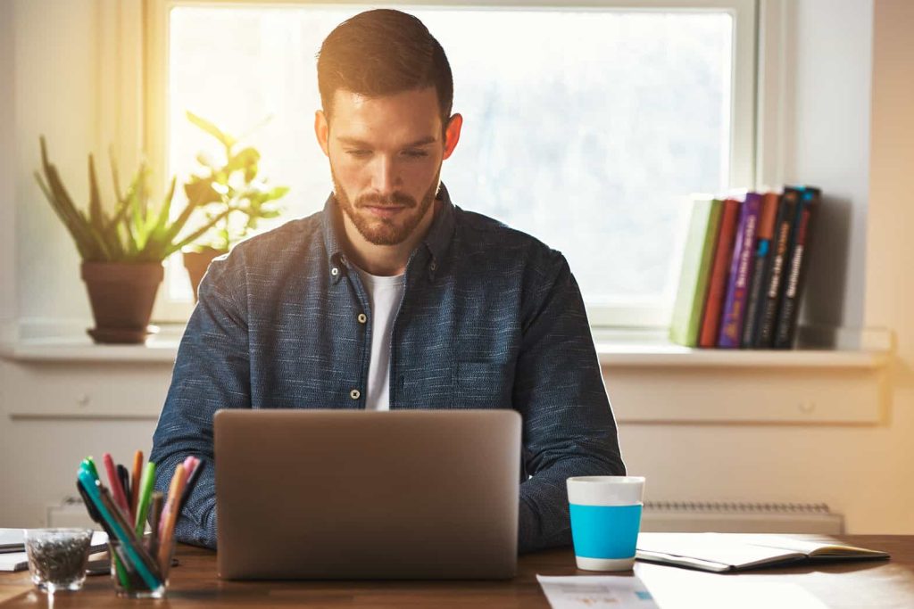 A bearded man wearing a blue shirt sits at a wooden table working on a laptop. There is a coffee cup, colorful pens in a holder, and a stack of books on the table. In the background, there is a window with sunlight streaming in and a potted plant on the sill.
