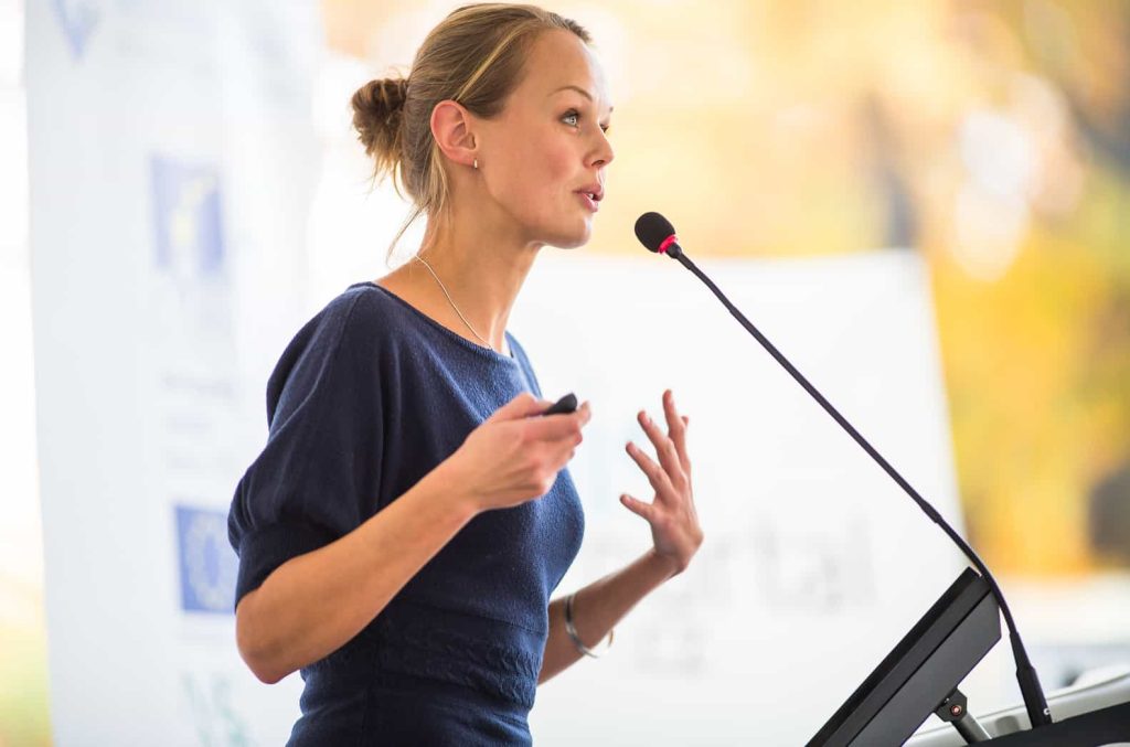 A woman with blonde hair tied back is speaking into a microphone on a stand. She is wearing a navy blue dress and is gesturing with her hands. She appears to be giving a presentation or speech in a brightly lit room with a blurred background.