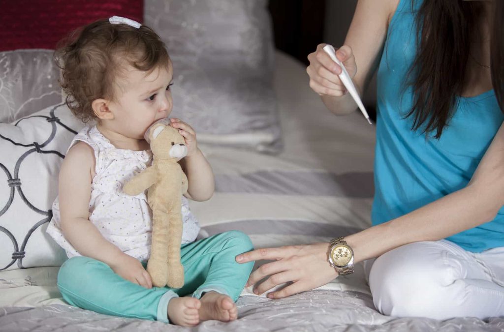 A young child sits on a bed, holding a stuffed toy and looking towards an adult who is holding a thermometer. The adult's hand and arm are extended towards the child.