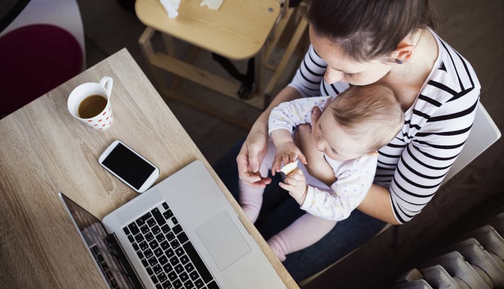 A woman is working on a laptop at a wooden table while holding a baby in her lap. The baby is playing with a small object. On the table, there is a smartphone and a cup of coffee. The focus is on the mother multitasking between work and childcare.