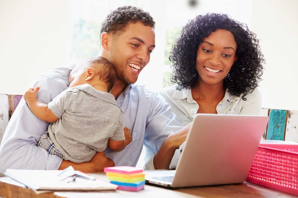 A smiling man holds a baby while looking at a laptop screen shared with a smiling woman. They sit at a table with a notebook, colorful sticky notes, and a pink basket. Sunlight streams in from a window behind them.