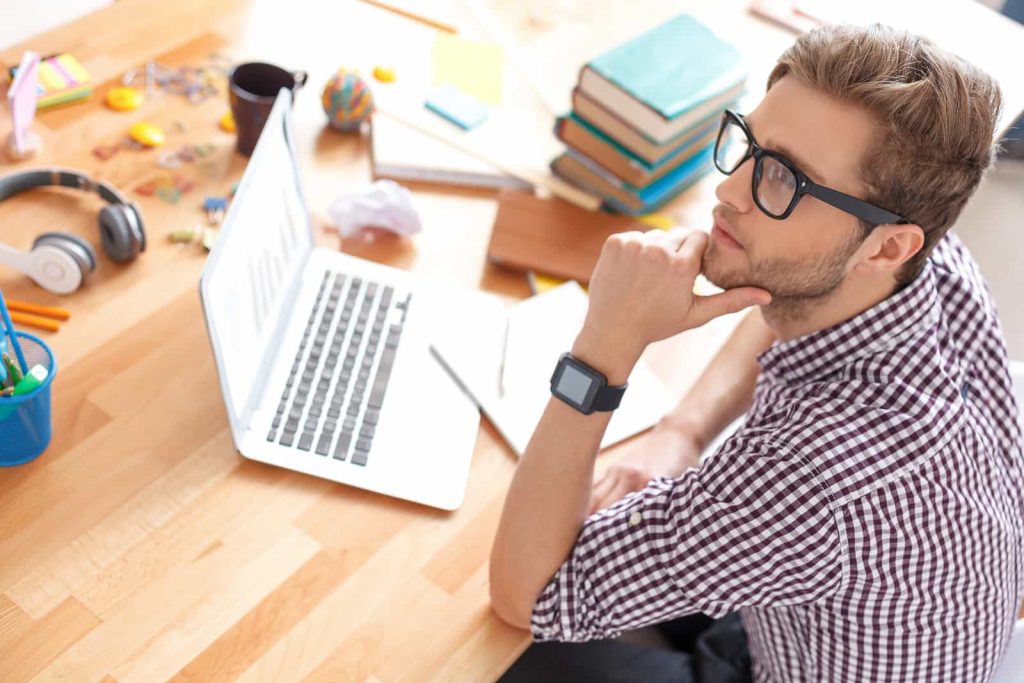 A man wearing glasses and a checkered shirt sits at a wooden desk, looking thoughtfully to the side with his hand on his chin. In front of him is an open laptop, surrounded by various items including books, headphones, and office supplies.