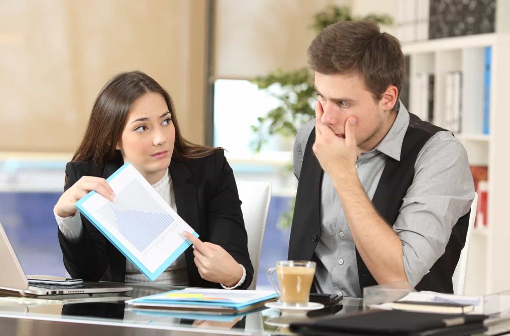 A woman in business attire is showing a document with a graph to a man, who appears worried and is touching his face. They are sitting at a desk with a laptop, papers, and a cup of coffee.