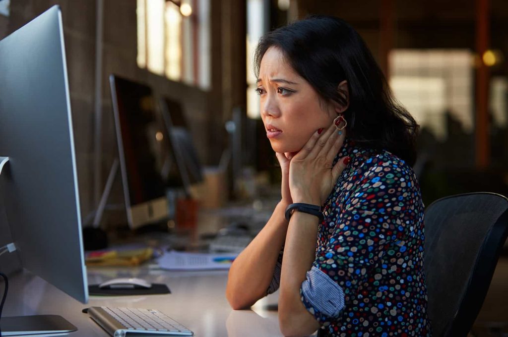A woman sits at a desk in front of a computer screen, looking worried and resting her hands on her neck. She is wearing a patterned blouse and a fitness tracker. The background shows an office environment with other desks and computers.