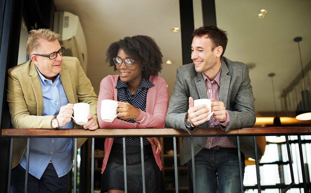 Three people, two men and one woman, stand at a railing holding coffee mugs and smiling. They are casually dressed and appear to be engaged in a friendly conversation in what looks like a modern indoor setting.