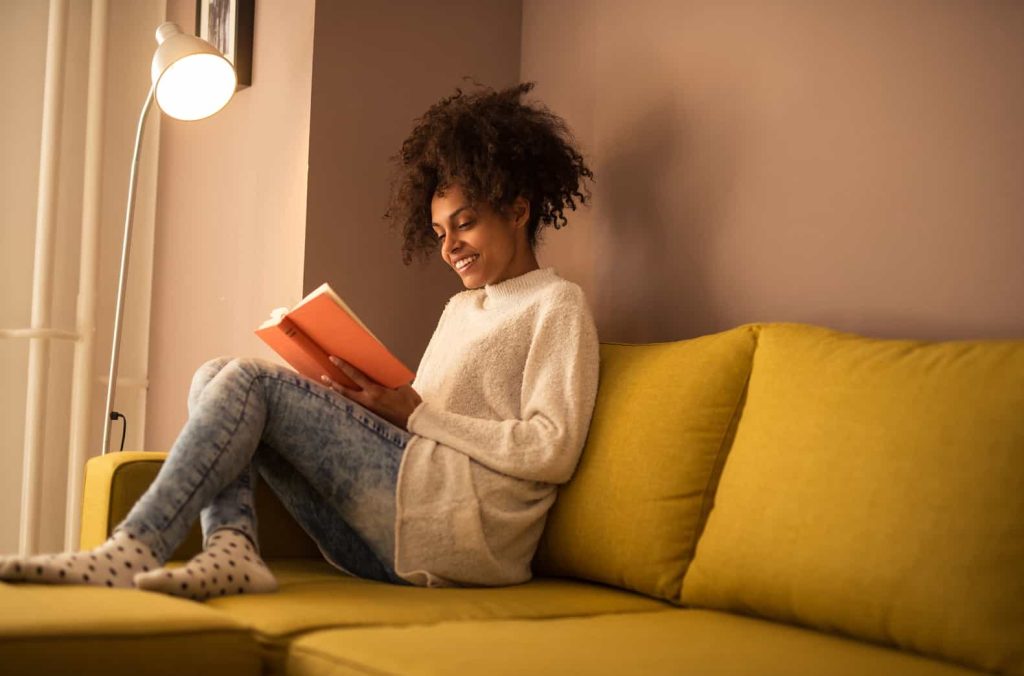 A person with curly hair, wearing a cozy sweater and jeans, is sitting on a yellow couch, reading a book. There is a floor lamp next to the couch, providing warm light. The background includes a softly colored wall, creating a comfortable, relaxed atmosphere.