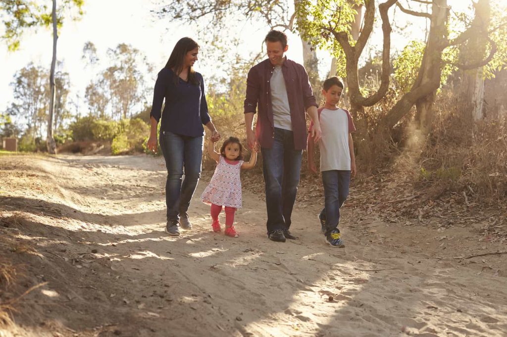 A family of four, including two adults and two children, walks hand-in-hand down a sunlit dirt path surrounded by trees.