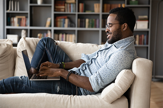 A smiling man lounging on a sofa while using a laptop, with bookshelves in the background. He is wearing glasses, a casual shirt, and jeans.