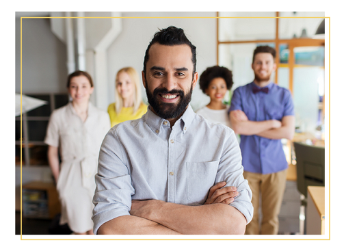 A confident man with a beard stands with his arms crossed in the foreground, smiling at the camera. Four colleagues, two women and two men, are blurred in the background, standing in a brightly lit office.